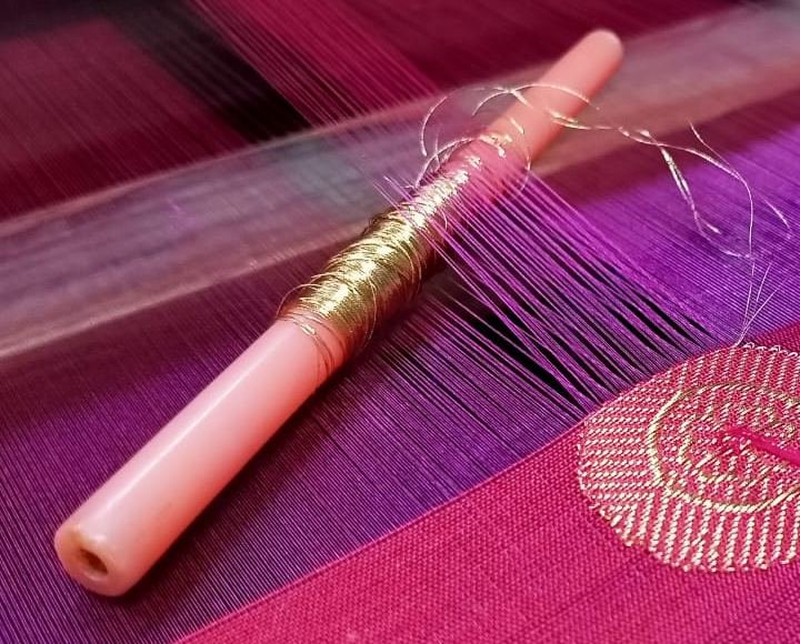 A bobbin of gold zari thread resting on magenta silk warp